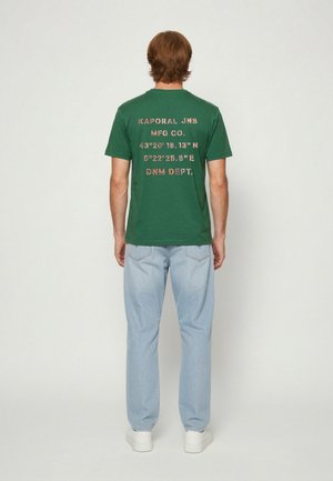 Homme debout, dos tourné, portant un t-shirt vert avec des coordonnées imprimées et un jean bleu clair sur un fond uni.
