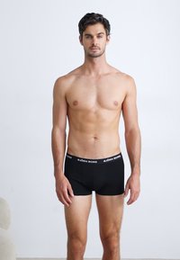 Schwarze Boxer-Shorts mit einem weißen „BJÖRN BORG“-Bund; taillierte Passform, glatte Textur und eine Länge bis zur Mitte des Oberschenkels. Model ist oberkörperfrei.