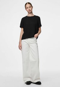 Blusa nera a maniche corte con scollatura rotonda abbinata a pantaloni bianchi a gamba larga. Il modello è in piedi con una mano in tasca, indossando scarpe nere.