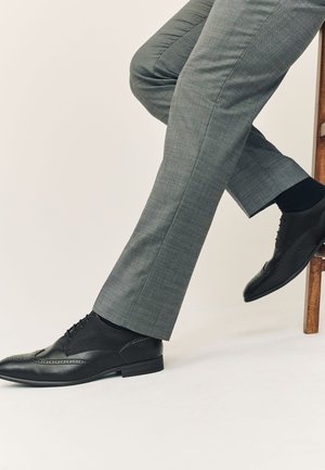 Homme portant un pantalon de costume gris et des chaussures brogues en cuir noir, assis avec une jambe croisée sur l’autre devant un fond uni.