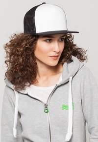 Crna i bijela snapback kapa sa strukturiranom prednjom stranom i mrežastim stražnjim dijelom, nosi se uz sivi duksericu s patentnim zatvaračem koja ima zeleni logo.
