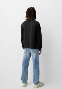 Osoba s dredovima nosi crnu majicu dugih rukava, svijetloplave traperice i bijele tenisice, stoji okrenuta bijelom zidu.