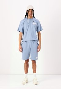 Maglietta di cotone azzurro chiaro con maniche corte, abbinata a pantaloni corti coordinati. Dettaglio del logo sulla maglietta, tessuto testurizzato e sneakers color crema.