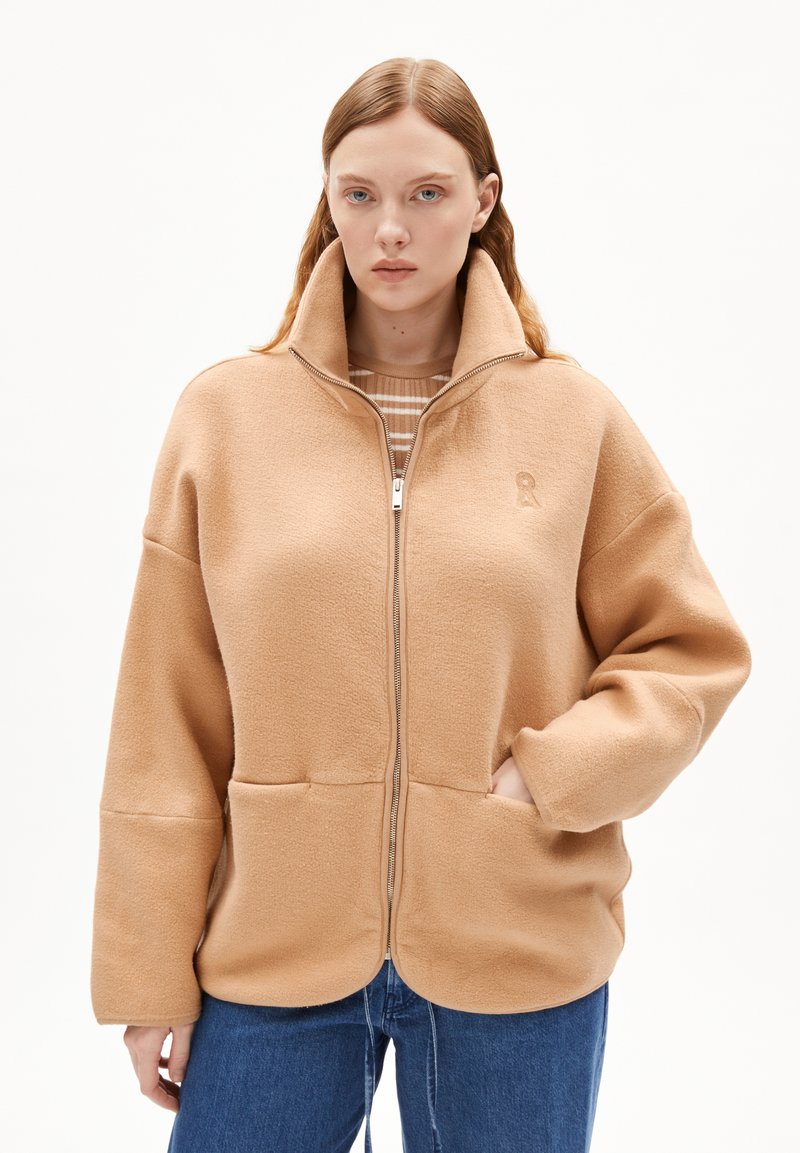 Beige fleecjas met hoge kraag en ritssluiting, voorzien van twee voorzakken en een subtiele logo-detail. Zachte textuur.