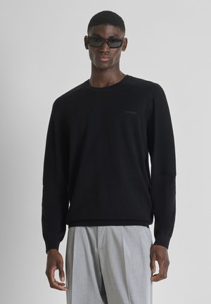 Homme portant un pull à manches longues noir avec un logo discret, un pantalon gris et des lunettes de soleil rectangulaires noires, debout devant un fond gris uni.