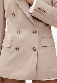 Bež dvoredni sako s pet smeđih gumba, dva prednja džepa i glatkom teksturom tkanine. Ima krojene reverje i krojeni kroj.