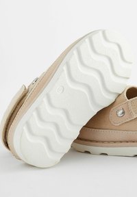 Chaussures beiges avec une tige en tissu texturé et une semelle en caoutchouc blanc présentant un motif ondulé. Accent en métal en forme de chat près de la sangle.