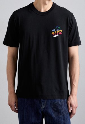 Man wearing black t-shirt with colorful abstract cyclist design on left chest, paired with dark blue denim jeans.
