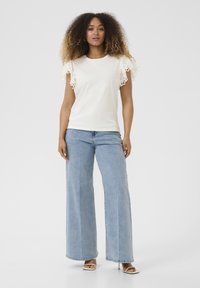 T-shirt blanc avec des détails en dentelle sur les manches, associé à un jean large taille haute bleu clair et des sandales à talons beiges.