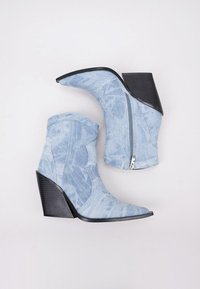 Botines de denim con puntera afilada, tacones angulados y cremallera lateral. Color azul claro con un patrón texturizado y desgastado, y acentos en el tacón de color negro.