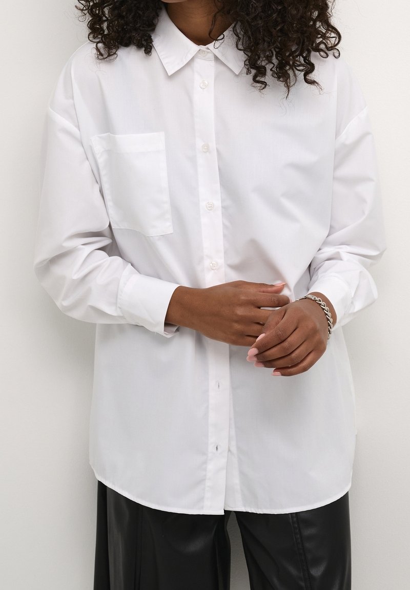 Personne aux cheveux bouclés portant une chemise blanche à boutons, un pantalon noir et un bracelet chaîne argenté, ajustant ses manches.
