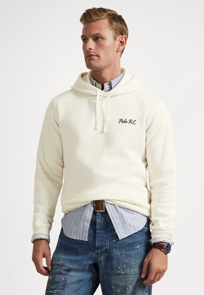 Muž v krémovej mikine s kapucňou cez modrú pruhovanú košeľu zapnutú na gombíky, ktorá je zastrčená v potrhaných džínsach s hnedom opaskom a hodinkami, pozerá doľava.