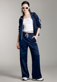 Marinblå träningsoverall med vita accentlinjer, bestående av en zip-up jacka och byxor med snörning. Modellen bär en vit crop top och solglasögon.