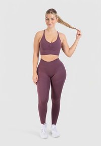 Lilanvärinen urheiluliivit ja korkeavyötäröiset leggingsit joustavasta kankaasta. Leggingseissä on sileä pinta ja istuva malli, täydentäen kokonaisuutta valkoisilla tennareilla.