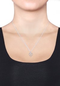 Silbernes herzförmiges Anhängerhalsband mit kleinen Steinen, das um den Hals einer Person über einem schwarzen Oberteil mit quadratischem Ausschnitt getragen wird.