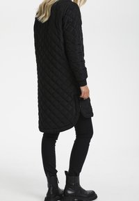 Schwarzer, gesteppter langer Mantel mit abgerundetem Saum, ausgestattet mit gerippten Bündchen und Fronttaschen, kombiniert mit schwarzen engen Hosen und knöchelhohen Stiefeln.