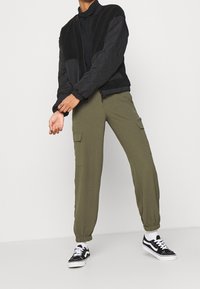 Veste bomber matelassée noire avec un dessus en polaire, associée à un pantalon cargo vert olive avec poches latérales et poignets élastiques, portée avec des baskets noires.