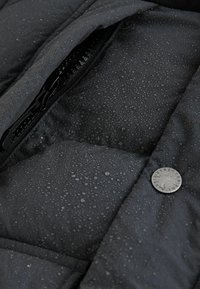 Black waterproof puffer jacket with visible water droplets, featuring a zippered pocket and a metal branded button on the exterior.