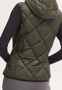 Pikowane oliwkowe bezrękawnik z kapturem noszone na czarnej bluzie z długim rękawem i szarych spodniach.