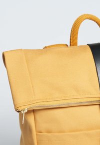 Zaino in tessuto giallo con design a roll-top, caratterizzato da un accento nero e una cerniera dorata. Superficie texturizzata con una robusta maniglia di trasporto.