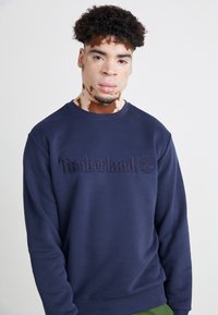Pulover din bumbac în nuanță de albastru marin, având un logo "Timberland" embosat și un grafic cu un copac. Designul include un guler rotund și manșete ribbate.