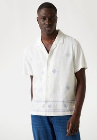 Short-sleeve white linen shirt featuring blue embroidery and a buttoned front. Patterns include geometric designs at the hem and chest.