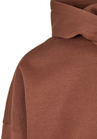 Gros plan du tissu marron d'un sweat à capuche montrant la texture et la capuche pliée sur un fond blanc uni.