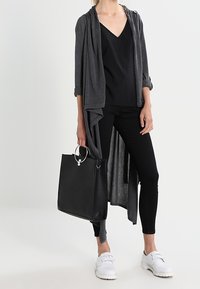 Larga chaqueta gris sobre una blusa negra de escote en V, combinada con leggings negros. Sosteniendo un bolso tote negro con un acento metálico y usando zapatos blancos.
