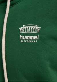 Tissu de sport vert avec logo hummel blanc présentant un design d'arche stylisée et un cordon beige sur le côté gauche.