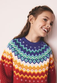 Barevný pletený svetr s kulatým výstřihem. Vzorem jsou cikcakové linie v červených, oranžových, žlutých, zelených a modrých odstínech.