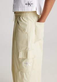 Ljusgula cargo-byxor med snörning i midjan, sidofickor och en bakficka med en vit broderad logotyp. Slät tygtextur.