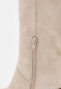 Beige suède kniehoge laars met een discrete zijrits en een gladde textuur, met gestikte details langs de naden.