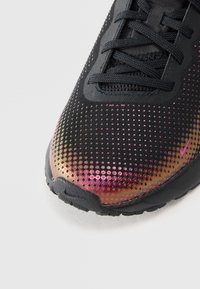 Μαύρο αθλητικό παπούτσι με υφασμάτινη επάνω επιφάνεια με υφή, που διαθέτει κλίμακα μεταλλικών κόκκινων έως χρυσών κουκκίδων στην άκρη, μαύρους κορδόνες και καουτσούκ σόλα.