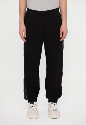 Black sweatpants with a relaxed fit, featuring an elastic waistband and side stripes. Fabric appears soft. Paired with light-colored sneakers.