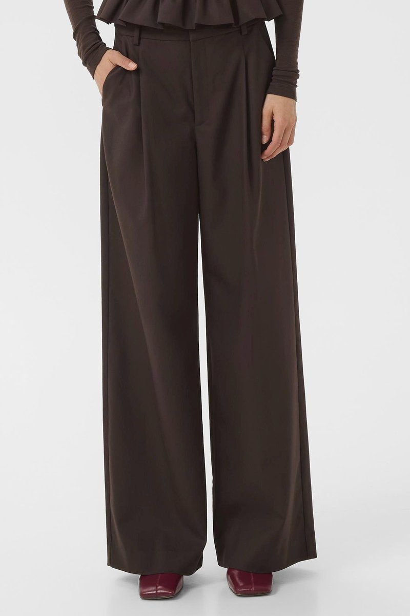Wide-leg trousers in dark brown fabric with a smooth texture, featuring pleats and side pockets. Styled with a layered top and burgundy shoes.