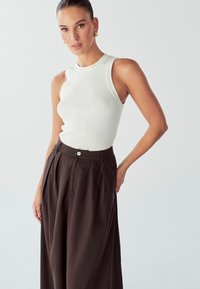 Weißes, geripptes Tanktop, kombiniert mit hochgeschnittenen, dunkelbraunen, weit geschnittenen Hosen mit Knopfdetails und Falten. Glatte Texturen und taillierter Schnitt.