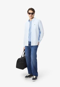 Lichtblauwe overhemd over een lichtblauw t-shirt, gecombineerd met donkere jeans en zwarte sneakers. Een zwarte tas wordt in één hand vastgehouden.