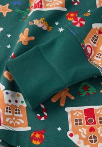 Vihreällä kankaalla on juhlallinen kuvio, johon kuuluu piparkakkutaloja, makeisia ja koristeita. Esitelty mansetti on sileä tekstuuriltaan ja siinä on värillistä ompelua.