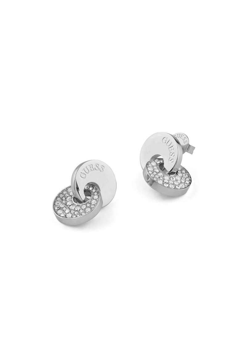 Silberfarbene Ohrringe mit einem kreisförmigen Design, das eine strukturierte Oberfläche und eingearbeitete klare Strasssteine aufweist. Das "GUESS"-Logo ist auf einem der Ohrringe sichtbar.