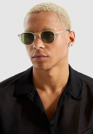 Ronde zonnebrillen met transparante monturen en groene lenzen, gedragen door een persoon in een zwart overhemd met kraag. Zilveren ooraccessoire zichtbaar.