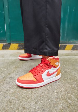 Persona con zapatillas altas en rojo, naranja y blanco, pantalones negros, de pie sobre cemento cerca de una puerta verde con cinta negra y amarilla.