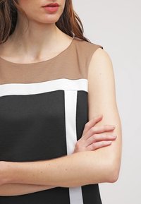 Haut sans manches avec une partie supérieure marron, une rayure blanche verticale et une partie inférieure noire. Texture lisse et silhouette ajustée.