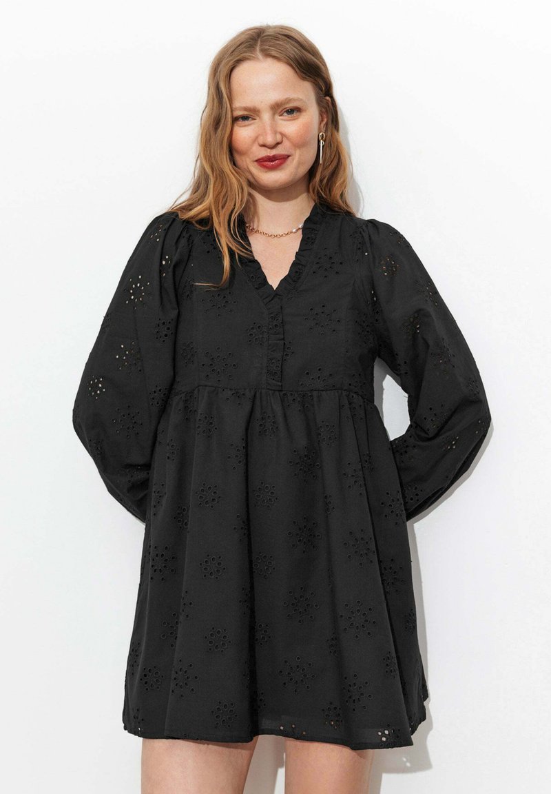 Femme souriante aux cheveux châtain clair portant une robe noire à manches longues avec des motifs floraux en œillets sur fond blanc.