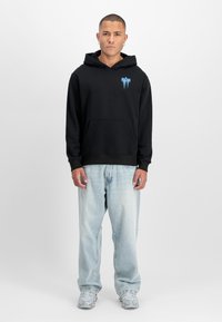 Svart hettegenser med frontlomme, med en blå grafisk logo på brystet, kombinert med lyseblå vide jeans og sølvfargede sneakers.