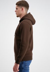 Brūns kapucēts džemperis ar priekšējo kabatu, ribotām aprocēm un gludu tekstūru, vilkts kopā ar ziliem džinsiem. Parādīts sānu skats.