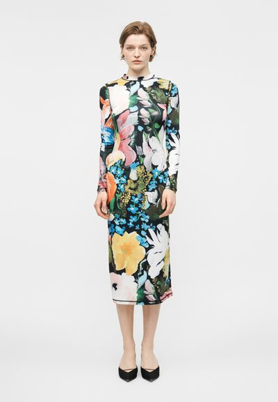 Femme debout, faisant face, portant une robe à manches longues, longueur genou, avec un grand imprimé floral coloré et des chaussures noires pointues.