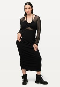 Schwarzes langärmeliges Kleid mit einem durchsichtigen Oberteil, gerafftem Design und taillierter Silhouette. Kombiniert mit schwarzen Schnür-Ankle Boots.