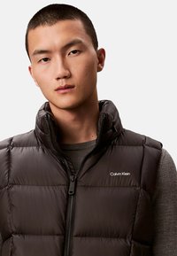 Czarna pikowana kamizelka z wysokim kołnierzem, zapinana na zamek błyskawiczny, z poziomym przeszyciem. Na piersi znajduje się biały logo Calvin Klein.