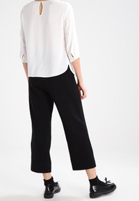 Vit blus med trekvartsärmar och nyckelhål på baksidan, kombinerad med svarta kapade byxor och polerade svarta skor.