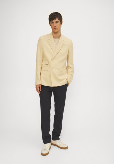 Beige dubbelzijdige blazer met twee voorkbeurzen, gecombineerd met marineblauwe broek en crèmekleurige sneakers. Soepele textuur, nauwsluitende pasvorm.